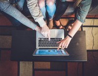 Drei Personen sitzen gemeinsam auf einem Sofa und zeigen gleichzeitig auf den Bildschirm eines Laptops, während sie an einem Tisch arbeiten.