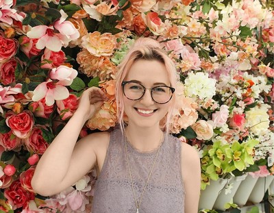 Eine junge Frau mit rosa Haaren und Brille lächelt vor einer üppigen Blumenwand mit Rosen, Orchideen und Pfingstrosen in zarten Pastelltönen.