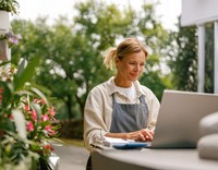 Eine Floristin in Schürze arbeitet lächelnd an ihrem Laptop im Freien, umgeben von Blumen und grüner Natur.